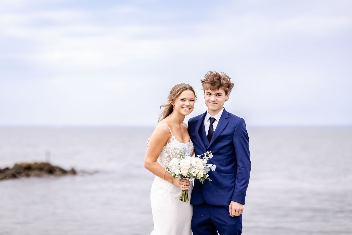 A couple in wedding attire walks along a misty beach with waves crashing behind them.