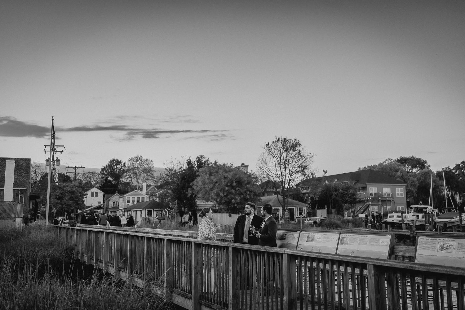 Black and white photo of people walking on a wooden bridge at dusk with a small town skyline in the background.