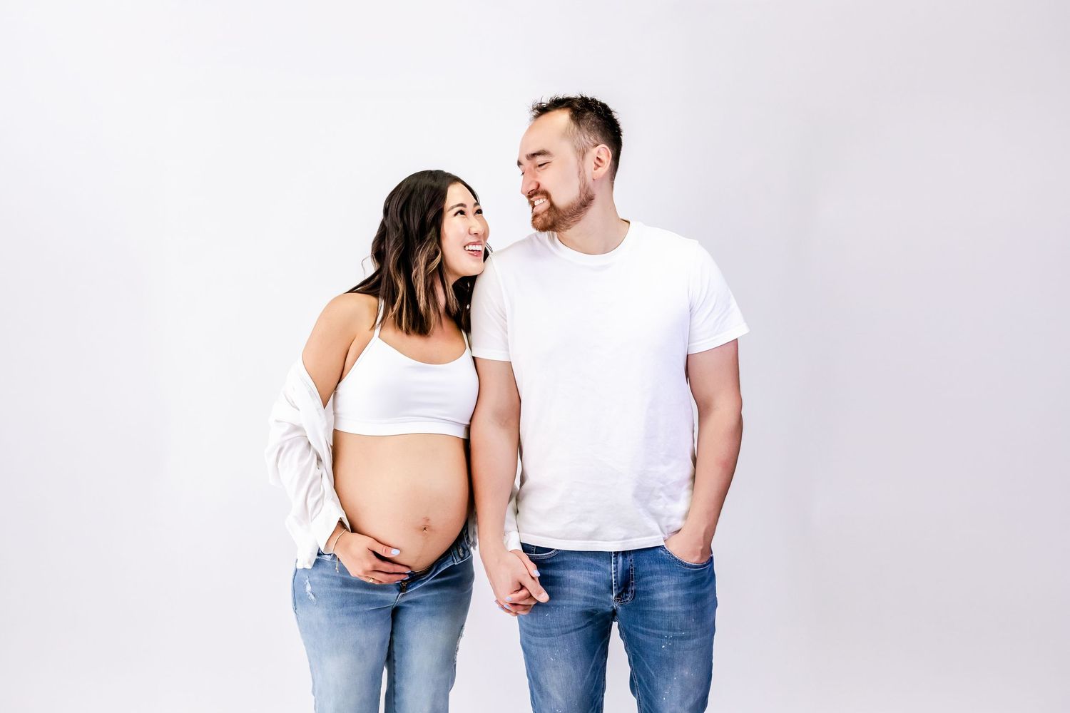 A couple in casual white and denim outfits pose together in a bright studio setting.