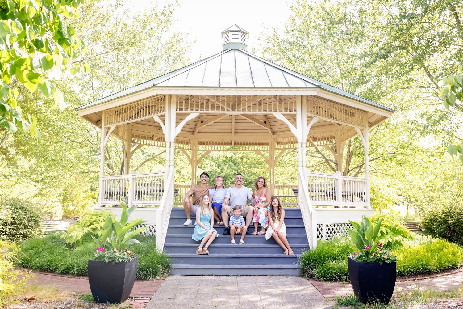 A white wooden gazebo in a garden setting with people gathered on the steps surrounded by lush greenery.