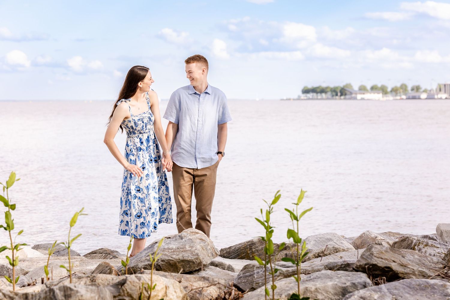 A couple stands together on rocky shoreline with water and blue sky in background, yellow flowers in foreground.