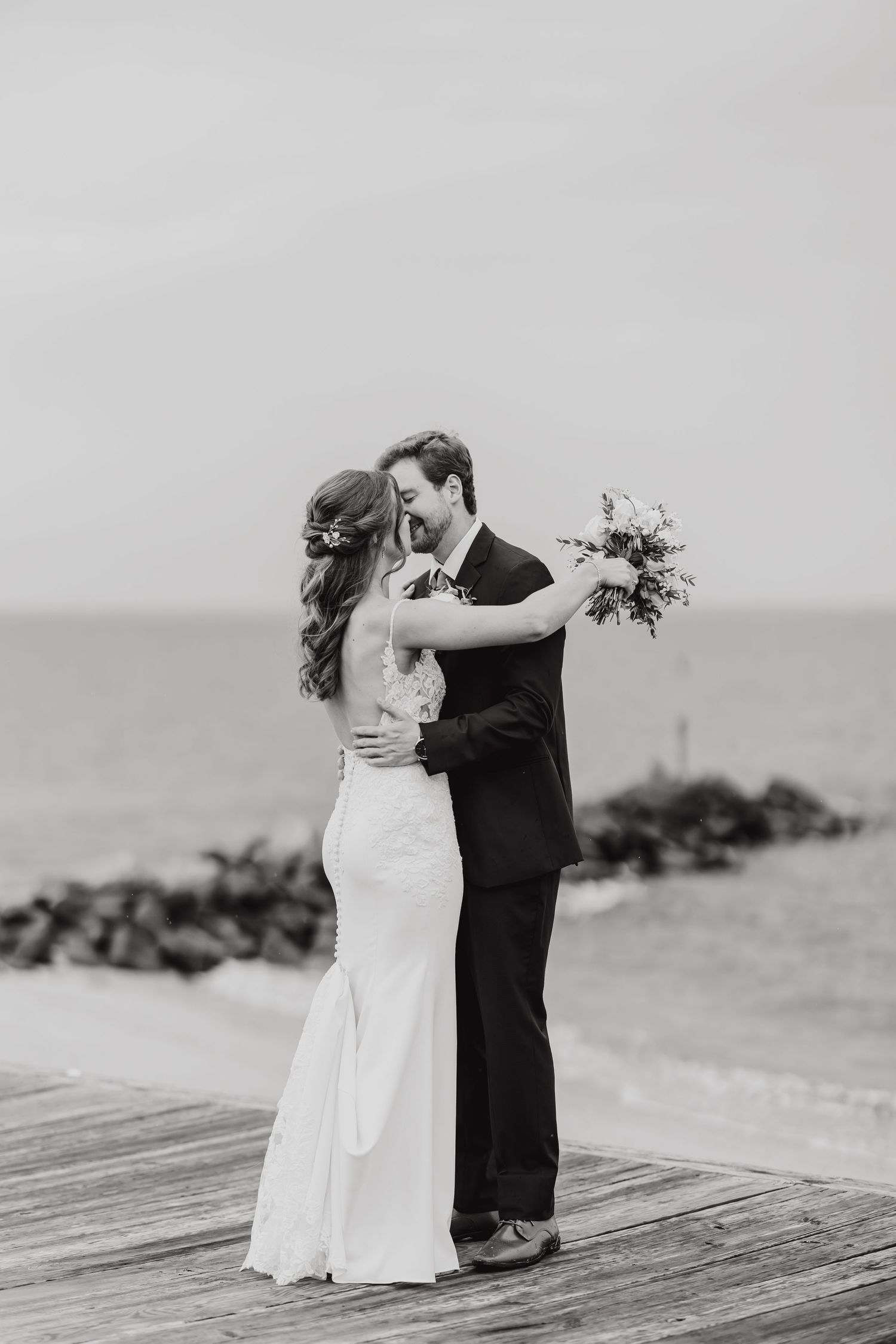 A black and white wedding photo of a couple embracing on a wooden pier by the ocean.