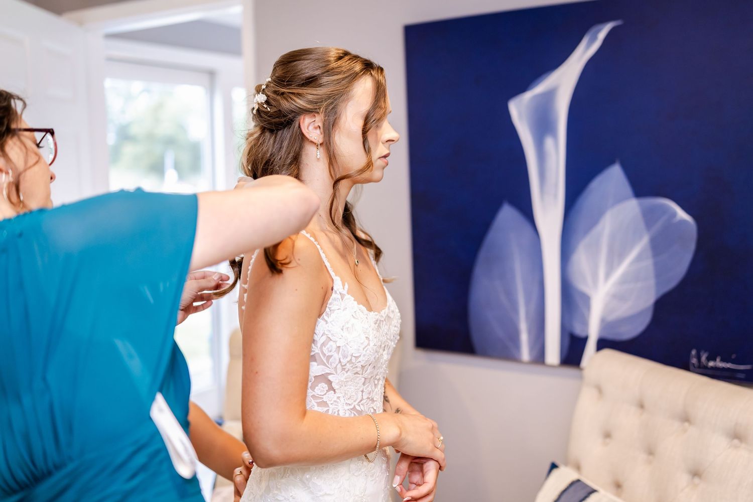 A bride in a white lace wedding dress stands in front of a blue wall with natural lighting.
