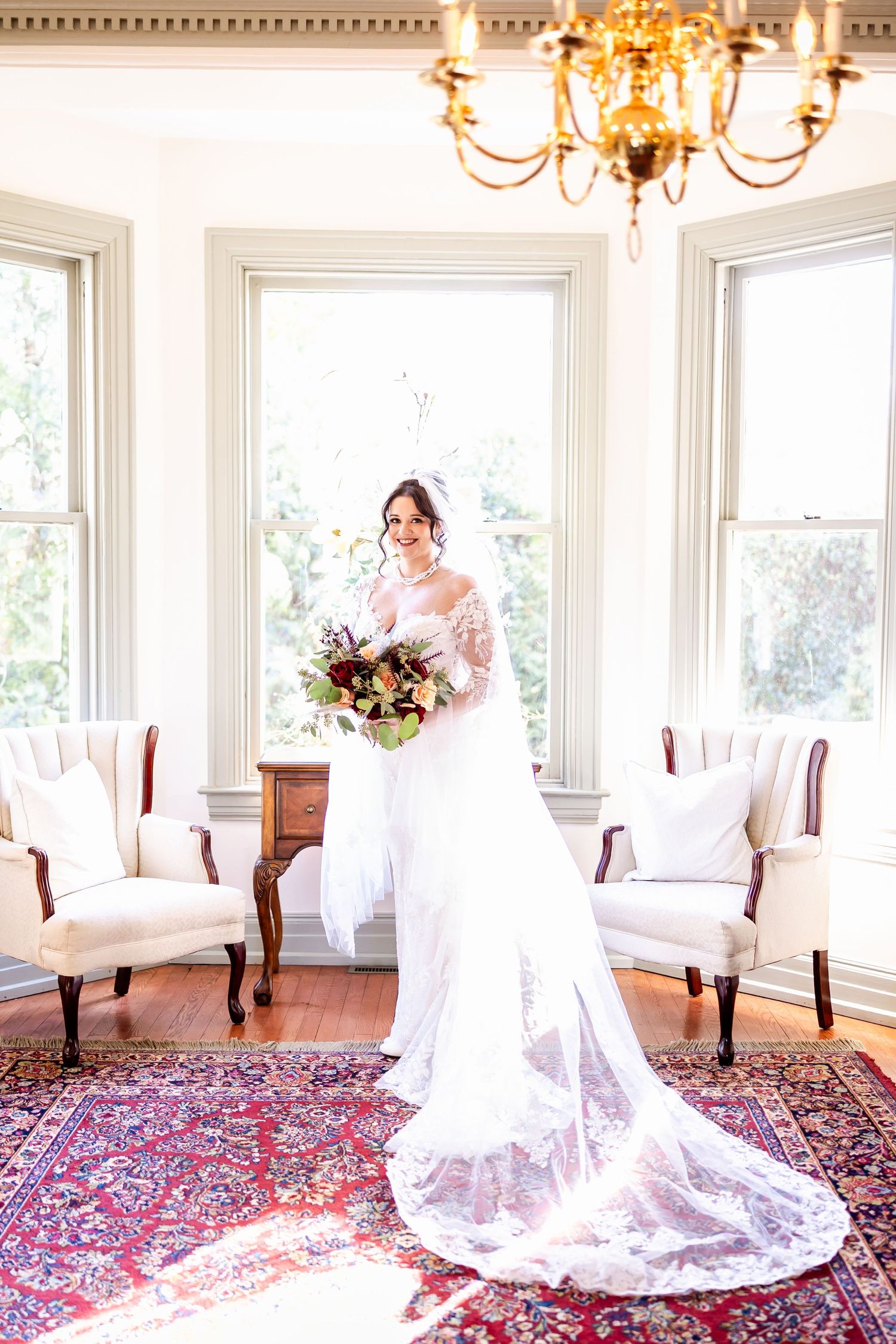 A flowing white wedding dress with a long train displayed in an elegant room with chandeliers and oriental rugs.