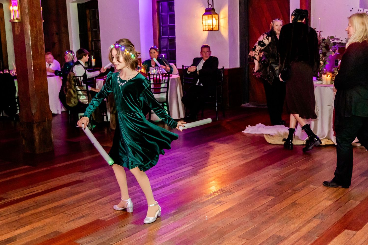 A guest in an emerald green dress dances energetically on a polished wooden dance floor at a wedding reception.