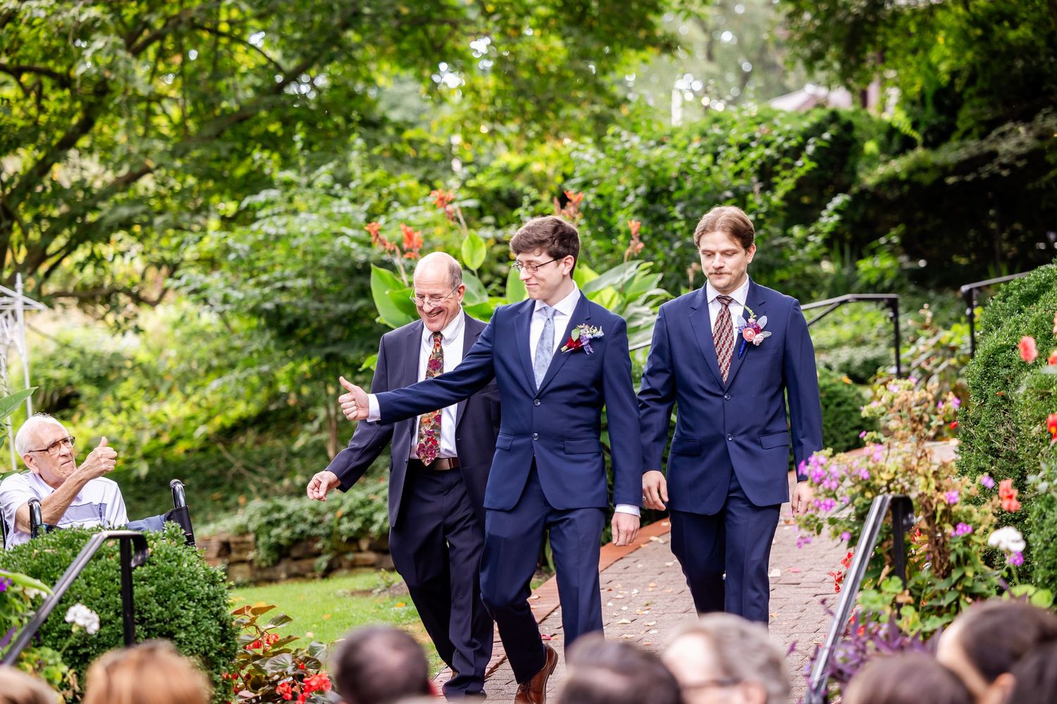 Three men in navy suits walk down a garden wedding aisle surrounded by lush greenery and flowers.
