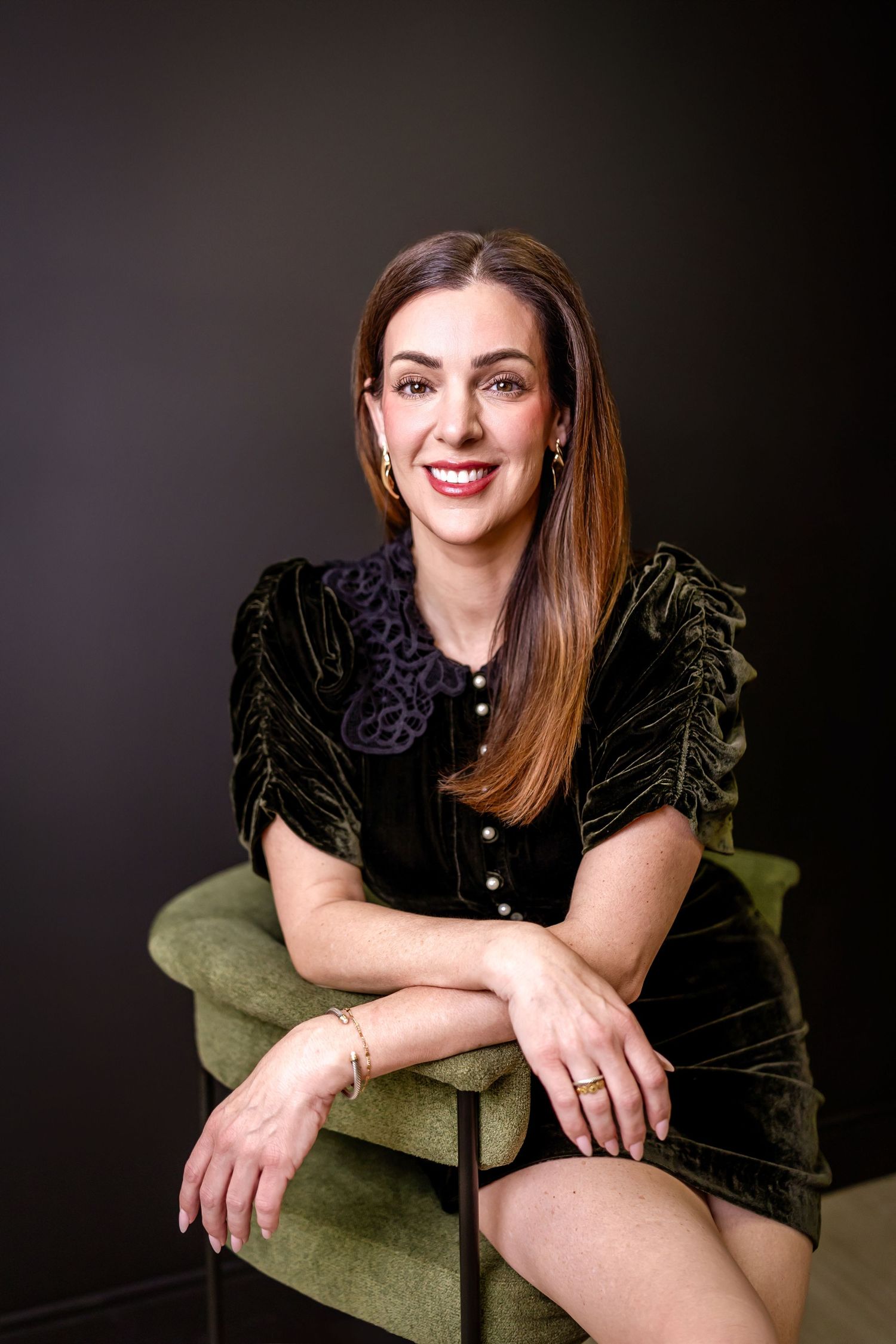 A smiling brunette woman in a black velvet dress sits elegantly on a green chair against a dark background.