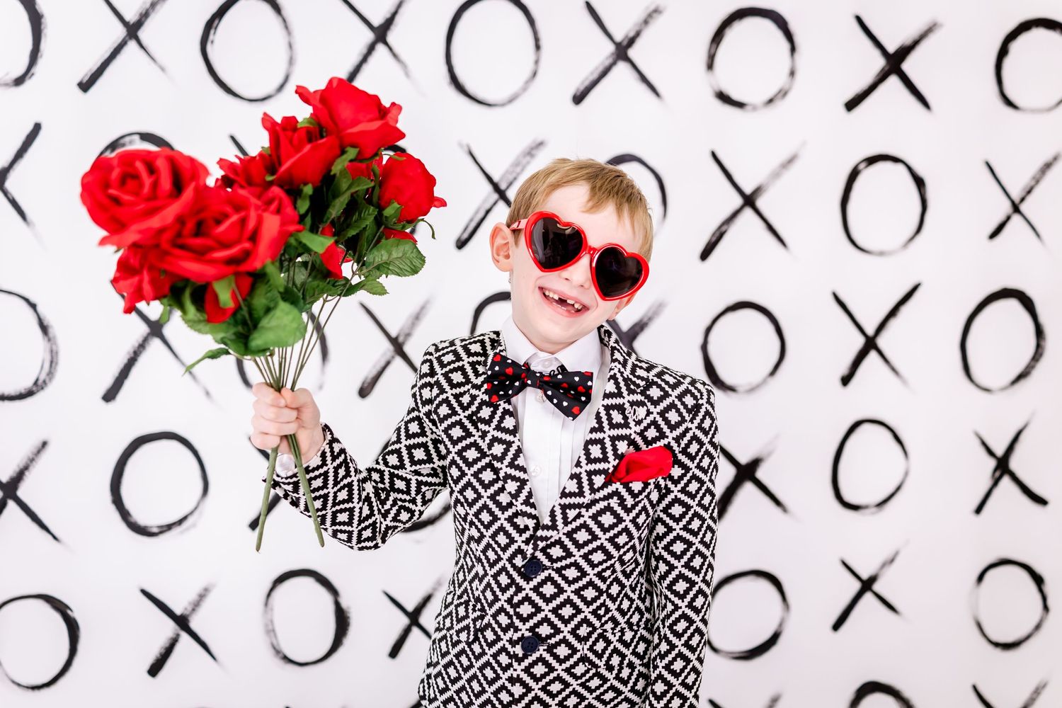 A figure wearing polka dot jacket and red heart sunglasses holds roses against a wall with XO patterns.