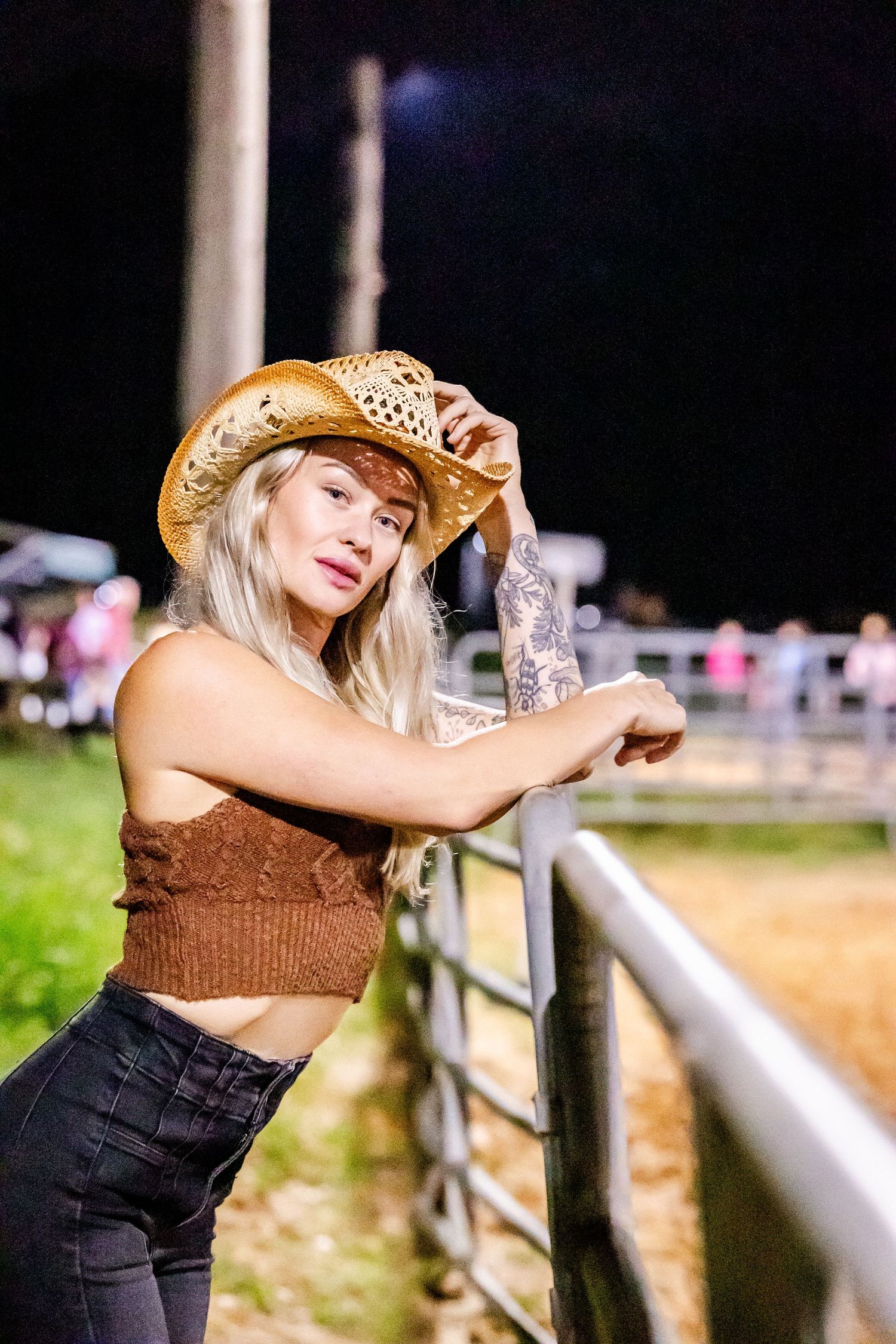Woman in cowboy hat leaning against white fence at rodeo arena during sunset.