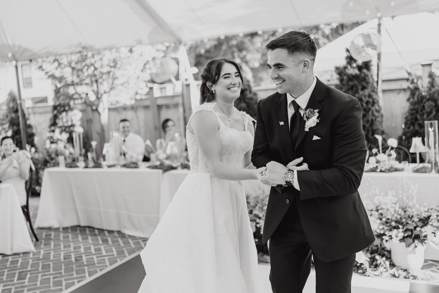 A black and white photo of a couple dancing together at an outdoor wedding reception with table settings visible.