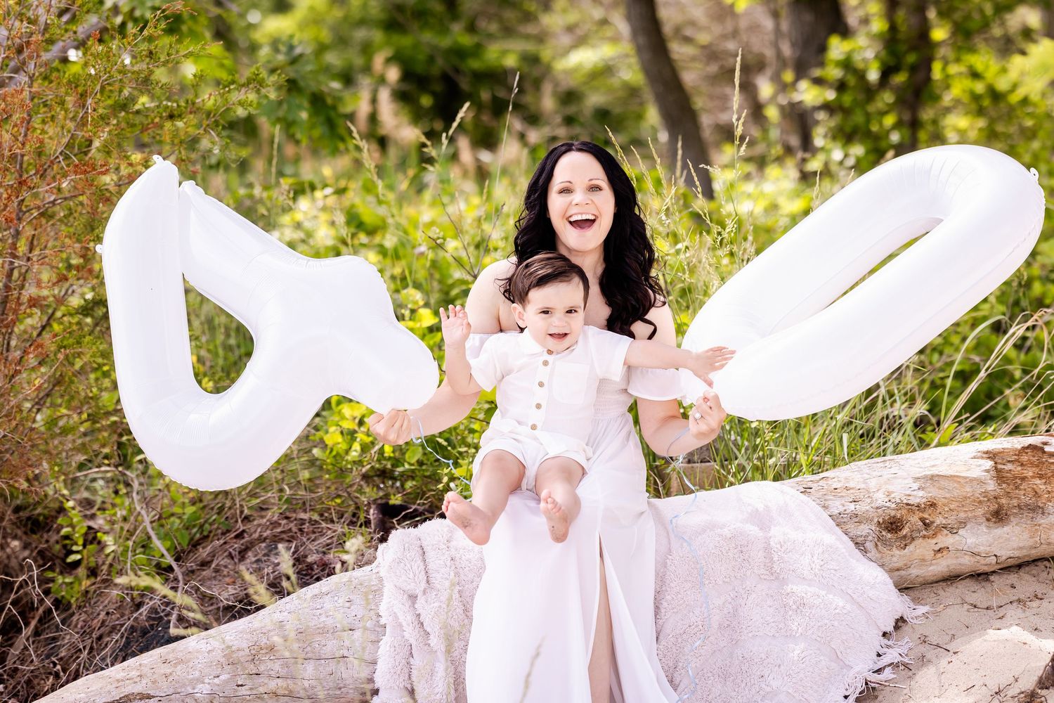 A mother and child in white outfits cuddle together during an outdoor photo session on a sunny day.
