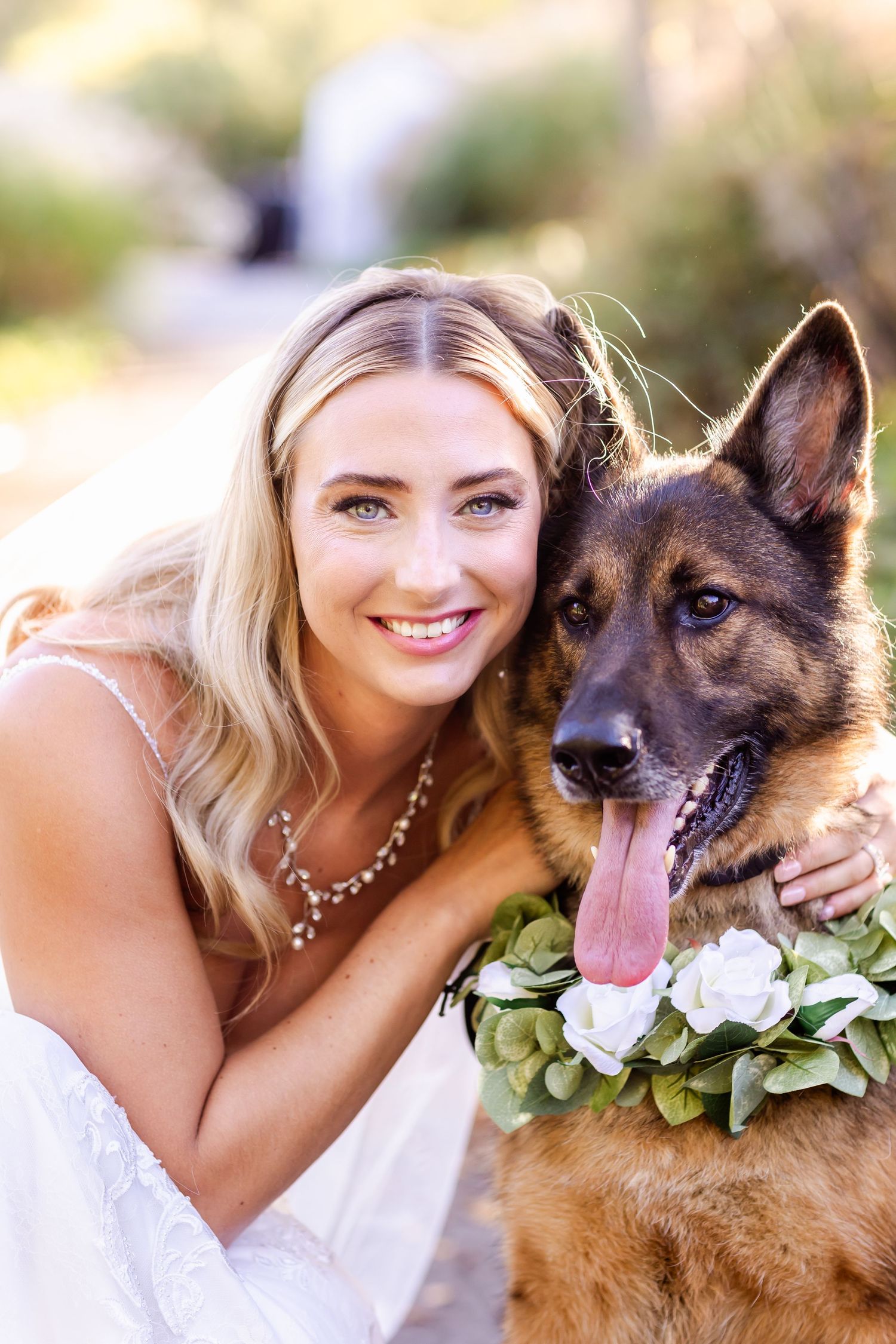 A German Shepherd wearing a floral collar poses with someone in a wedding dress during an outdoor photoshoot.
