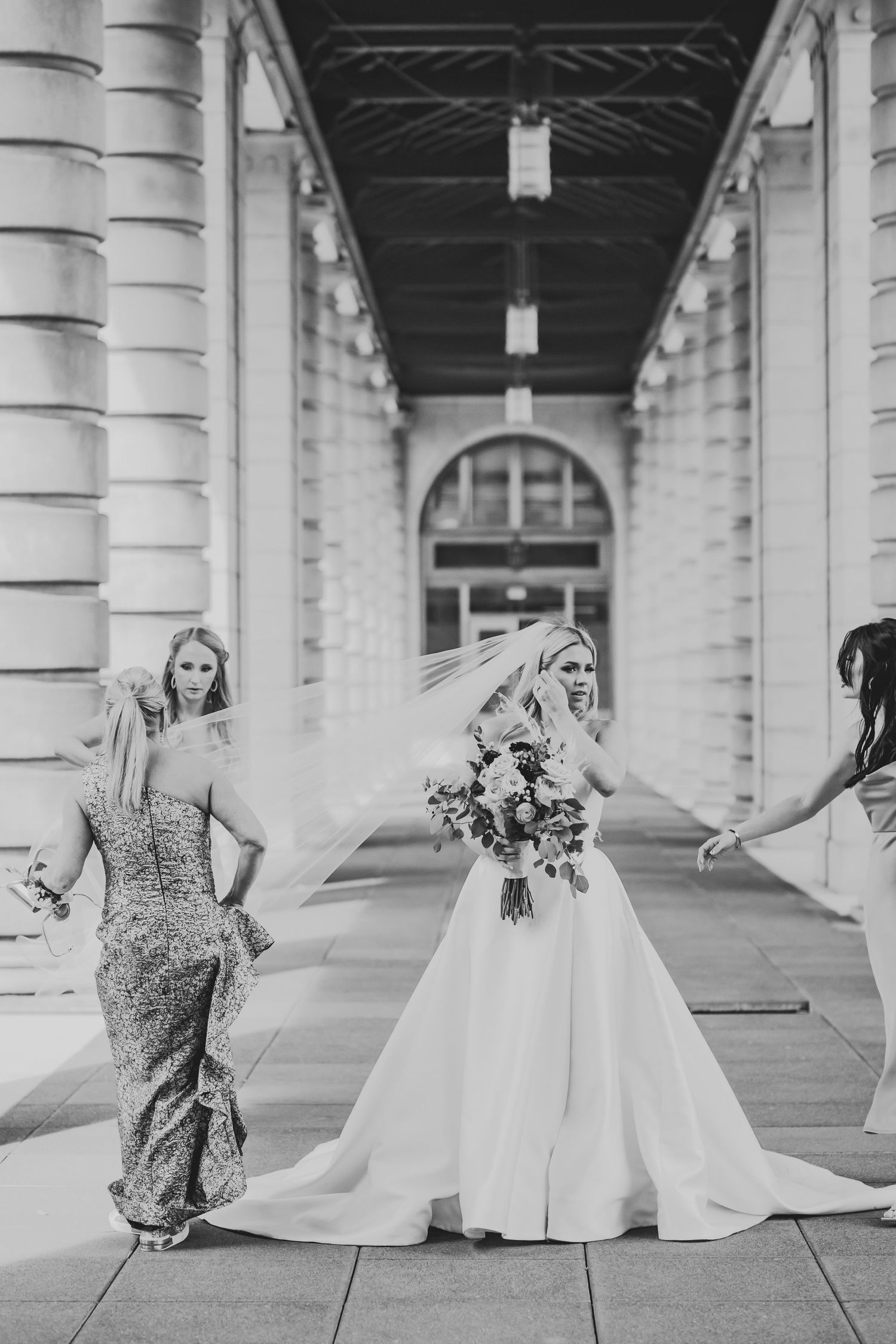 Black and white photo of three bridal party members walking under columned corridor with wedding dress and bouquet.