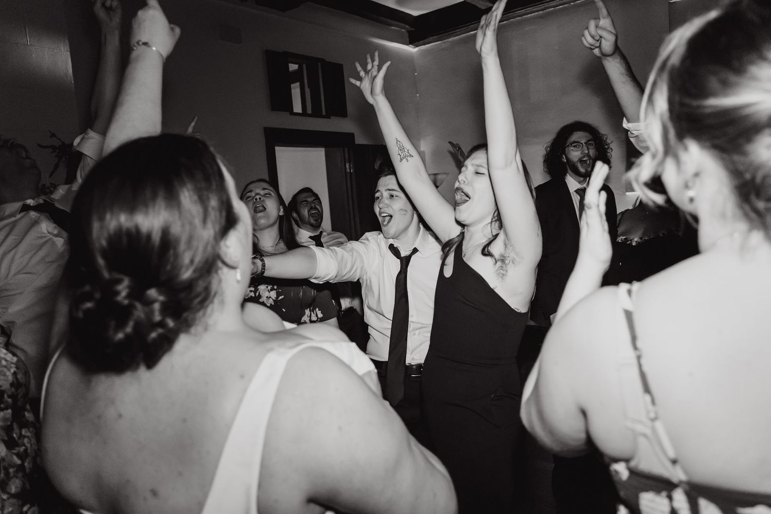 Black and white photo of wedding reception dance party with raised hands in celebration.