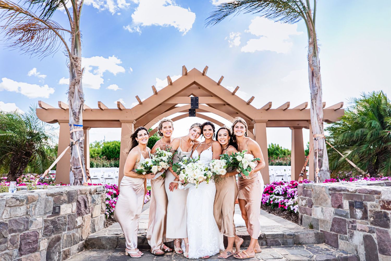 Wedding party poses under wooden pergola with palm trees and pink flowers in tropical setting.