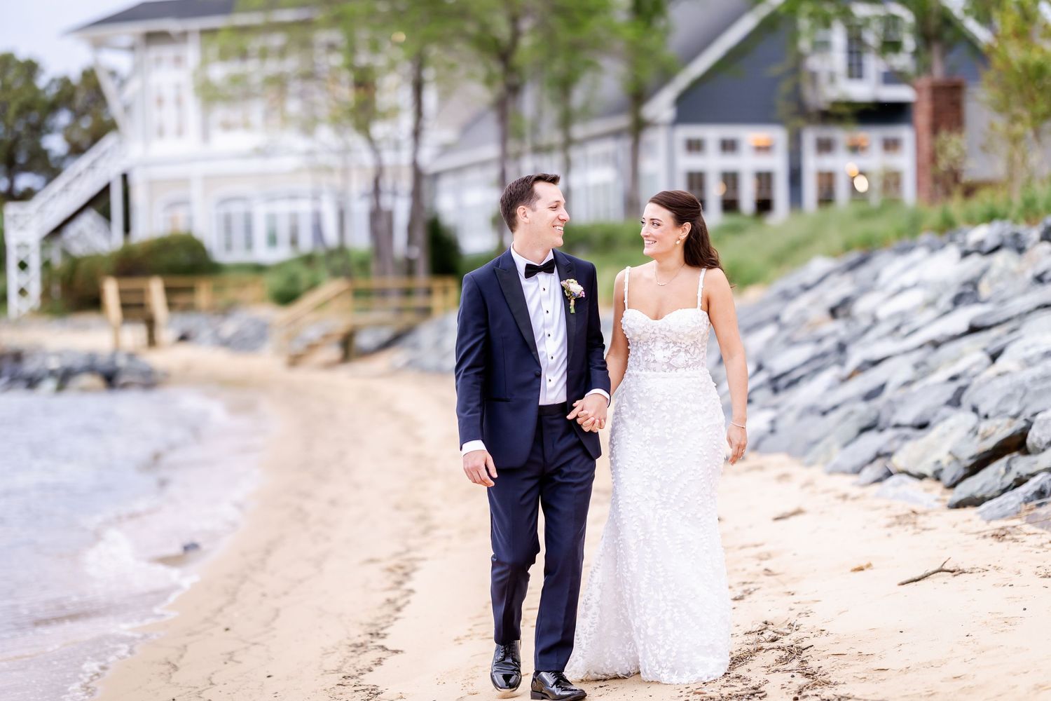 A couple in wedding attire walks along a rocky beach shoreline during sunset, creating romantic silhouettes.