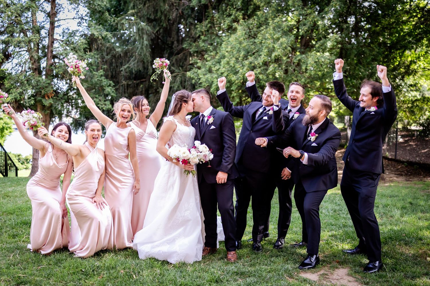Wedding party in pink dresses and dark suits poses playfully on grassy lawn.
