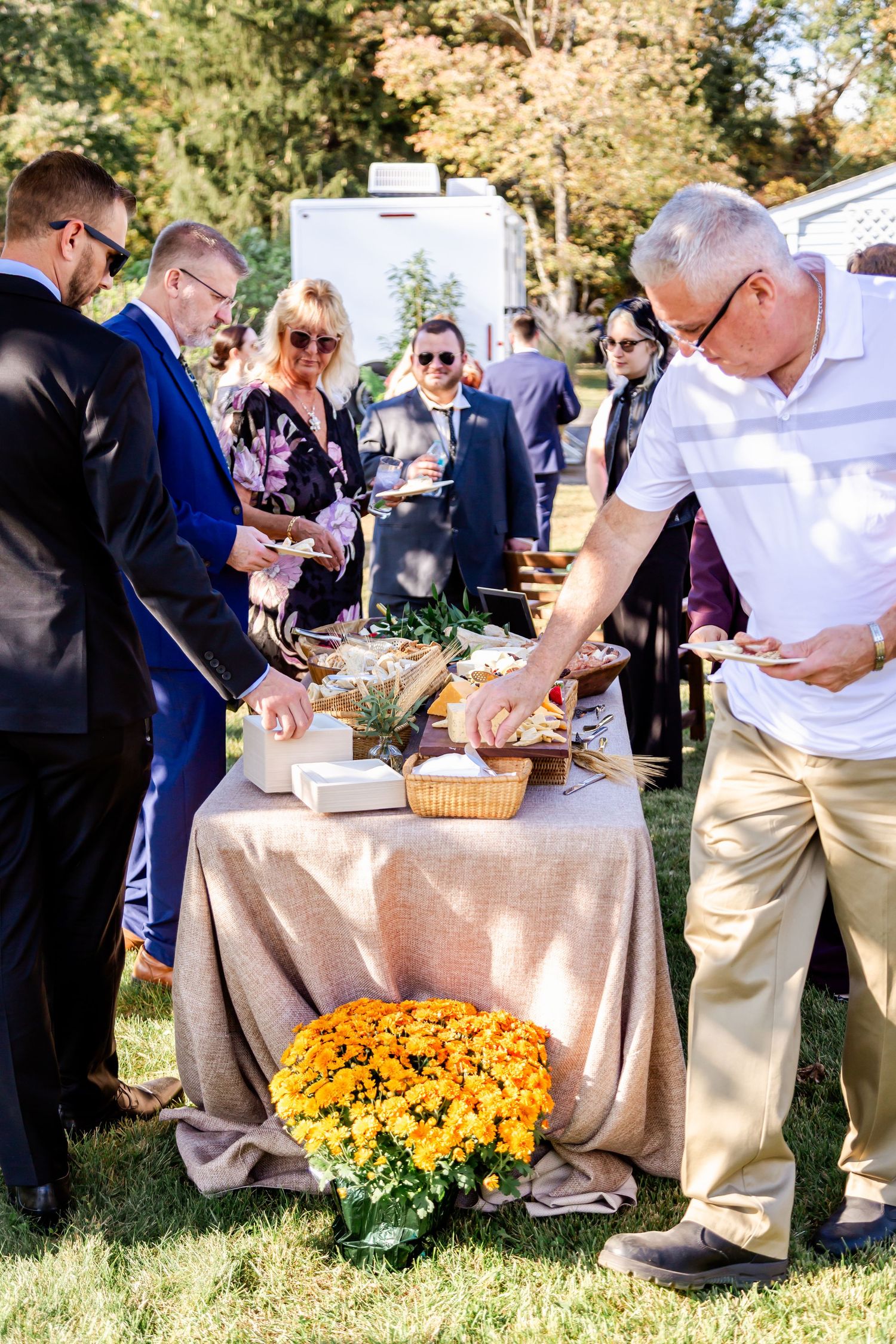 Wedding guests gather around a buffet table with yellow mums decorating the outdoor setup.