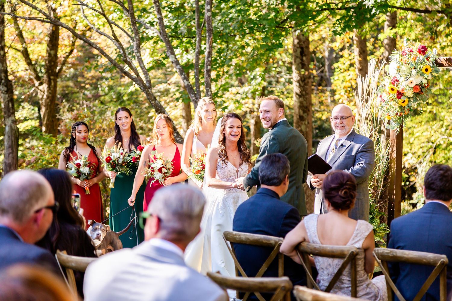 An outdoor wedding ceremony in a wooded setting with guests seated on wooden chairs.