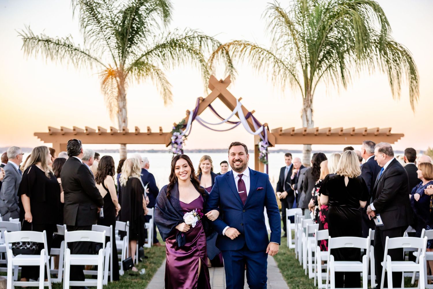Wedding party members walk down aisle at sunset outdoor ceremony with palm trees.