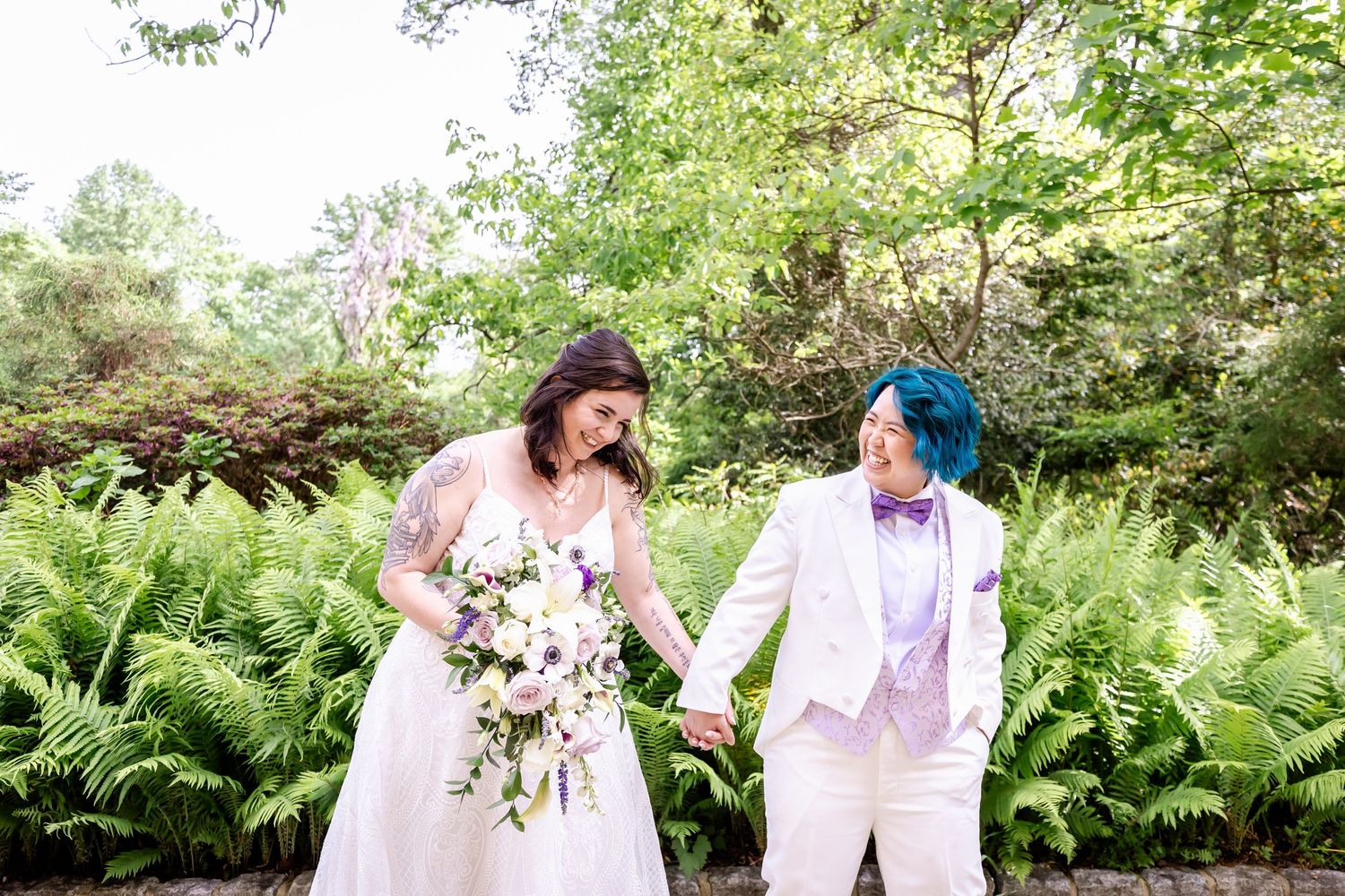 A happy couple shares a joyful moment in a garden surrounded by ferns, both wearing light colored attire.