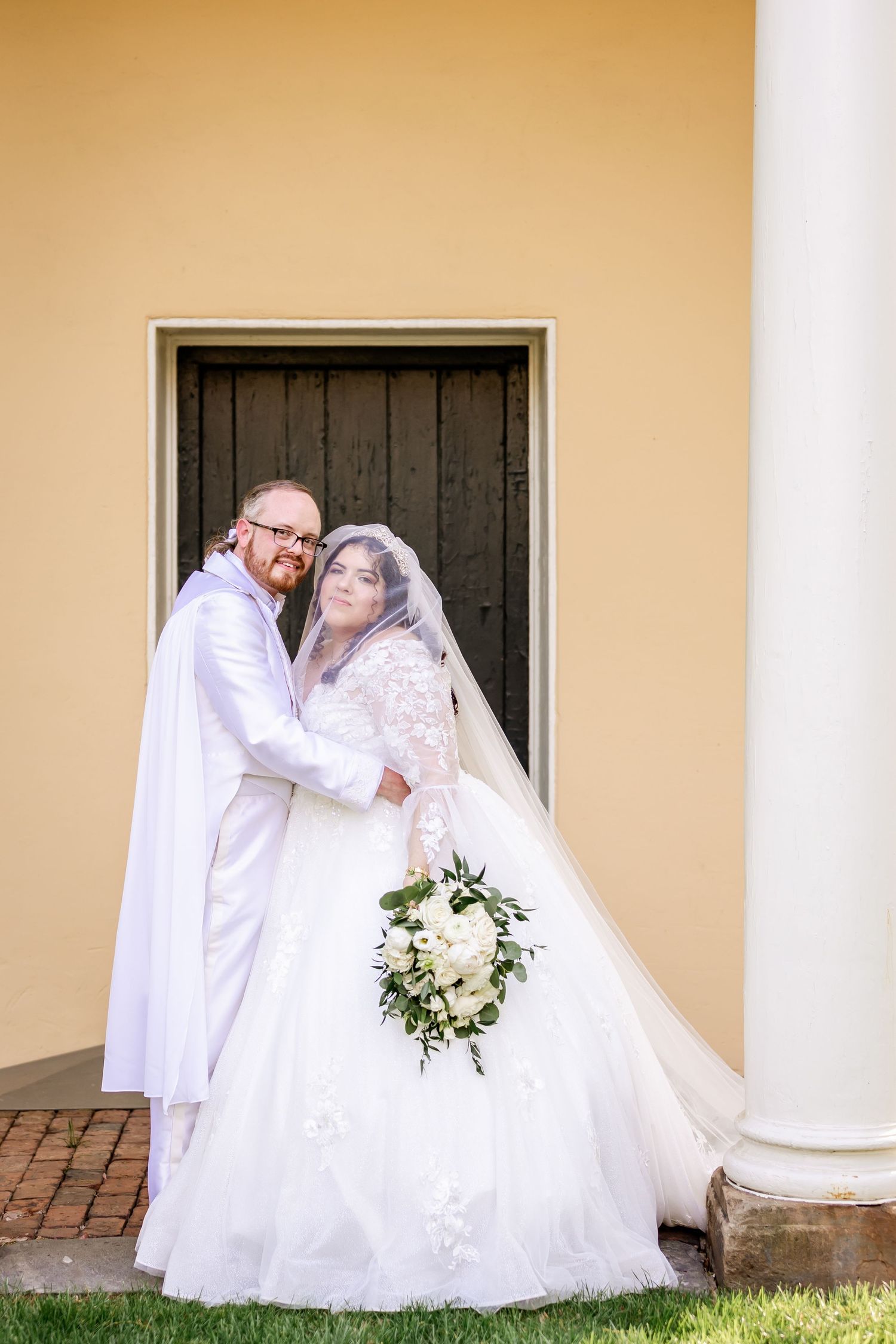 Loving embrace captured during wedding photoshoot against yellow colonial architecture.