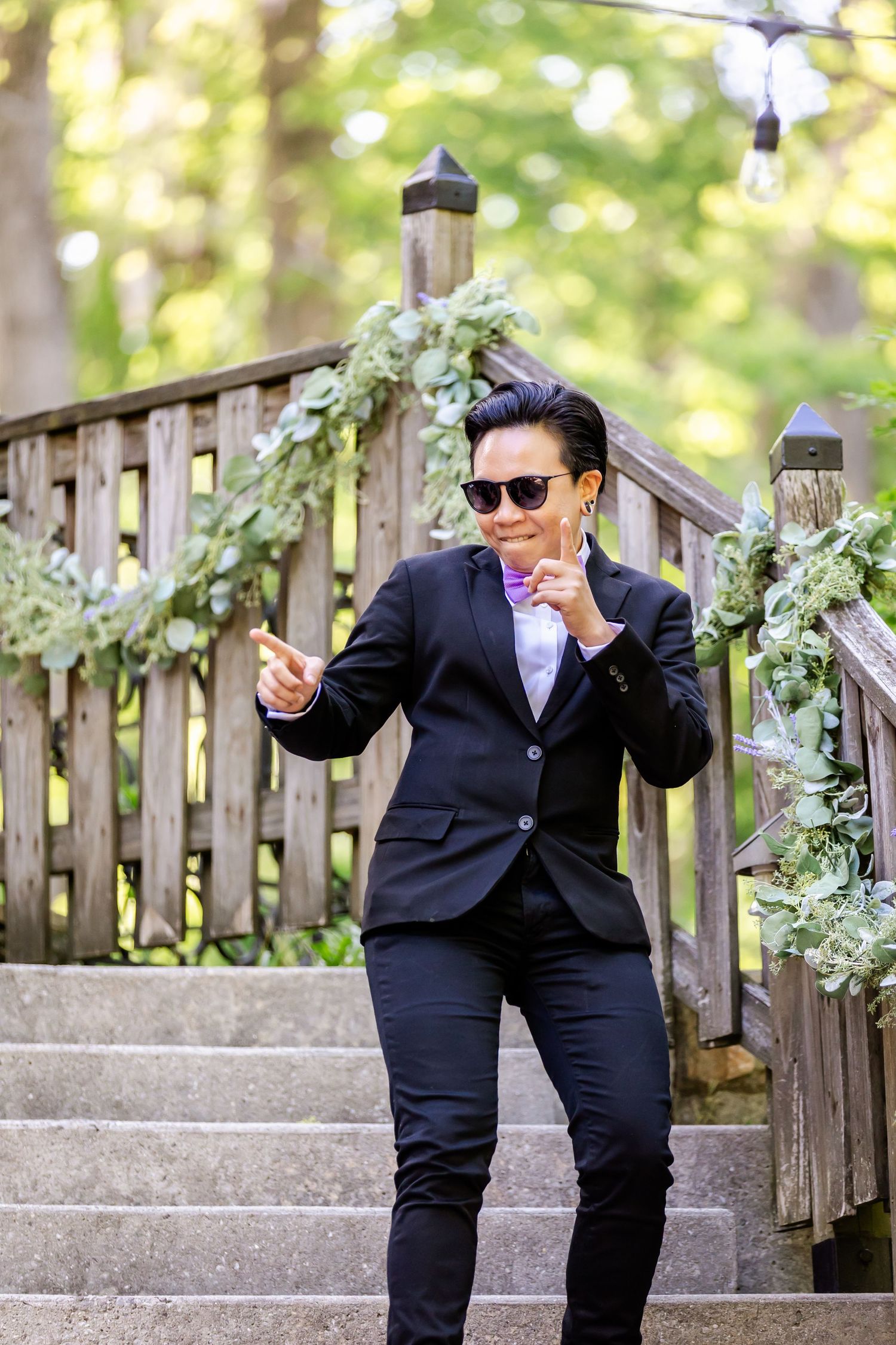 Person in dark suit strikes playful pose on outdoor staircase decorated with greenery.