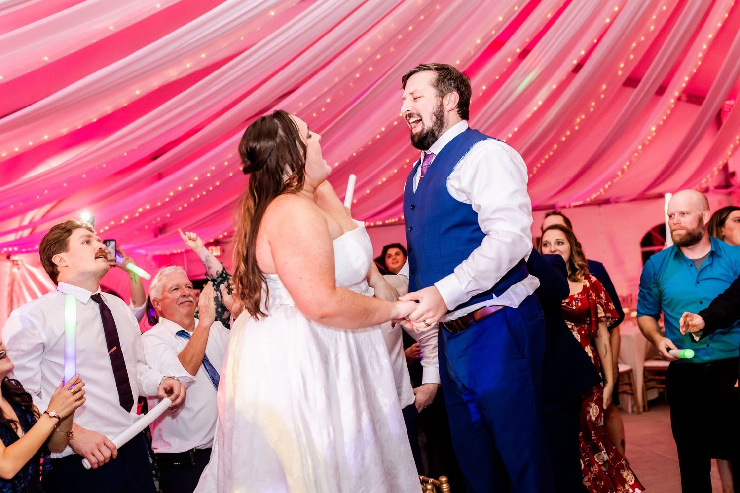 Couple dancing at their wedding reception under pink draped ceiling with guests cheering.