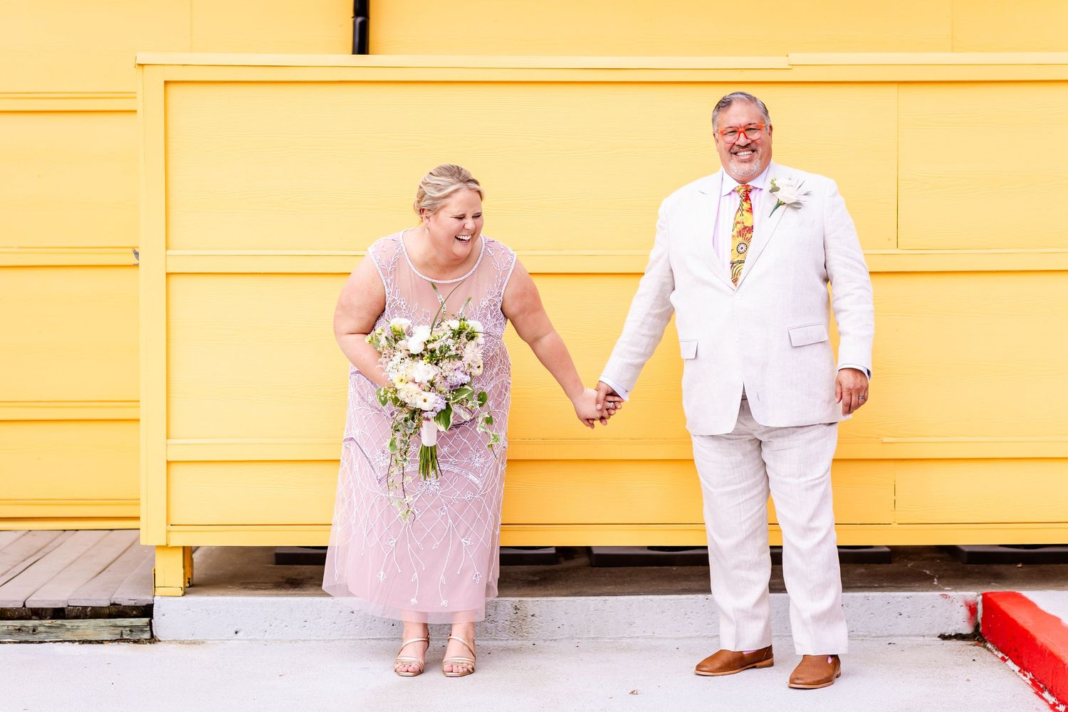 A couple holds hands while standing in front of a bright yellow wall in coordinated formal wear.
