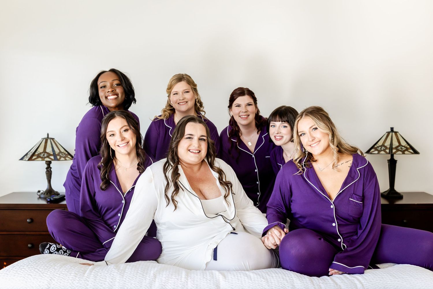 Wedding party in matching purple robes pose together on bed during pre-ceremony preparations.