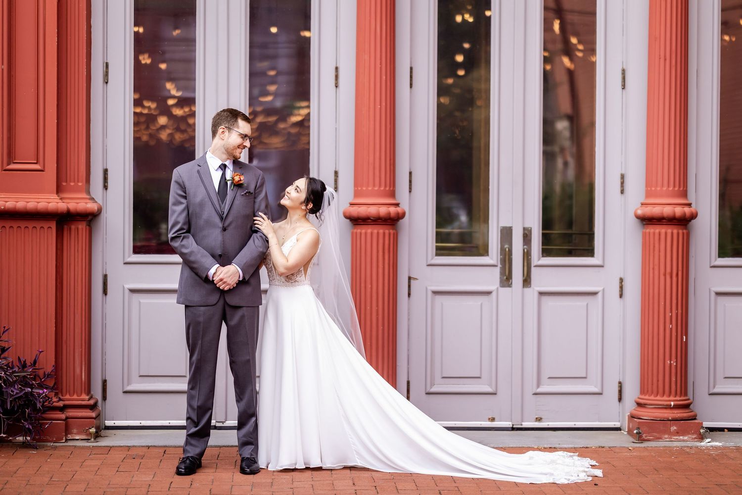 A romantic wedding couple poses in front of a red brick building with elegant gray doors and windows.