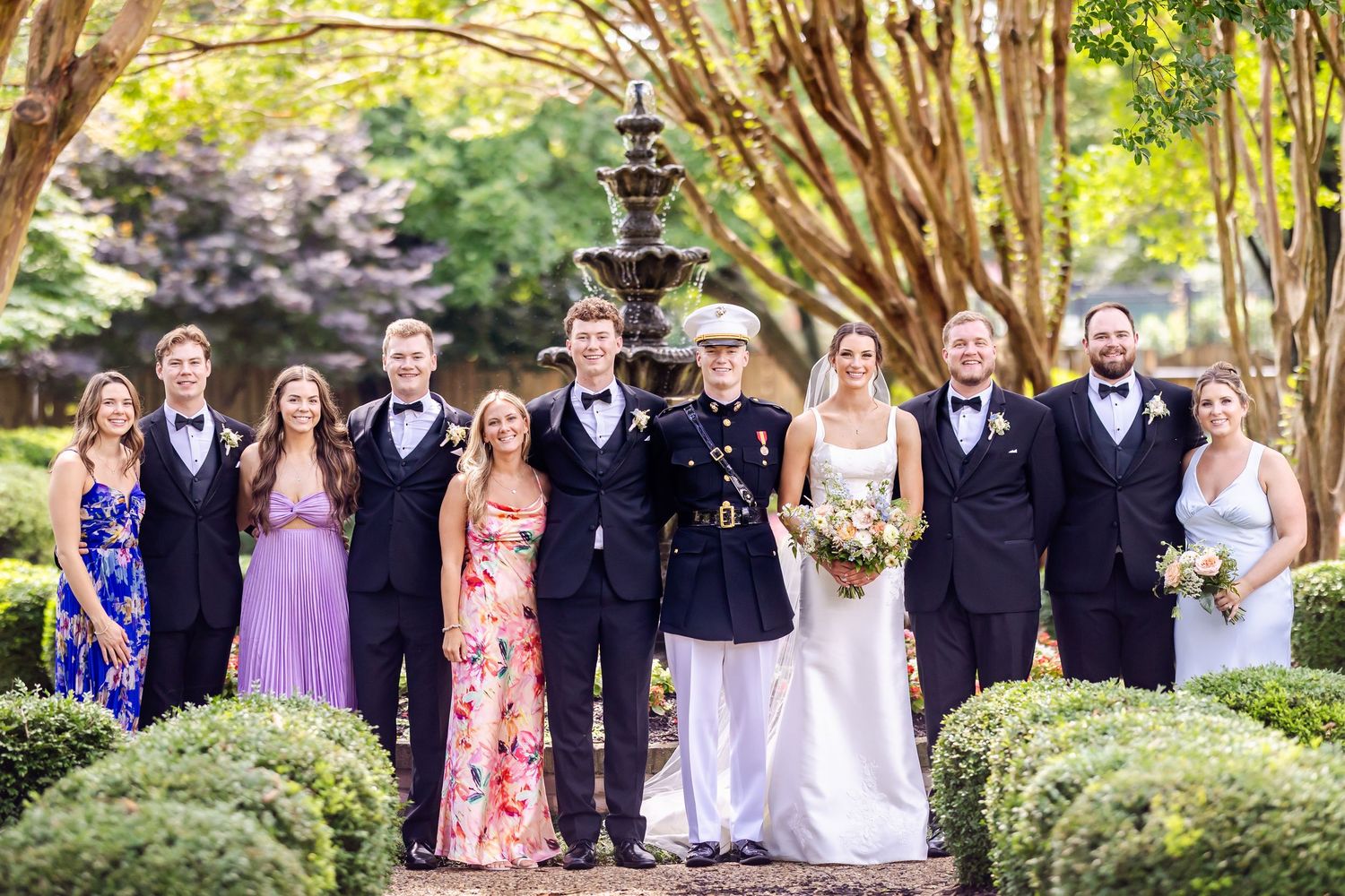 Wedding party in formal attire poses together on garden path with fountain backdrop.