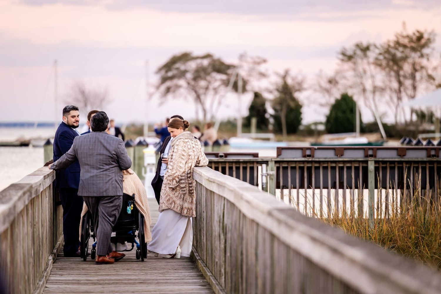 Group of people on a wooden boardwalk by the water at sunset, including someone in a wheelchair.