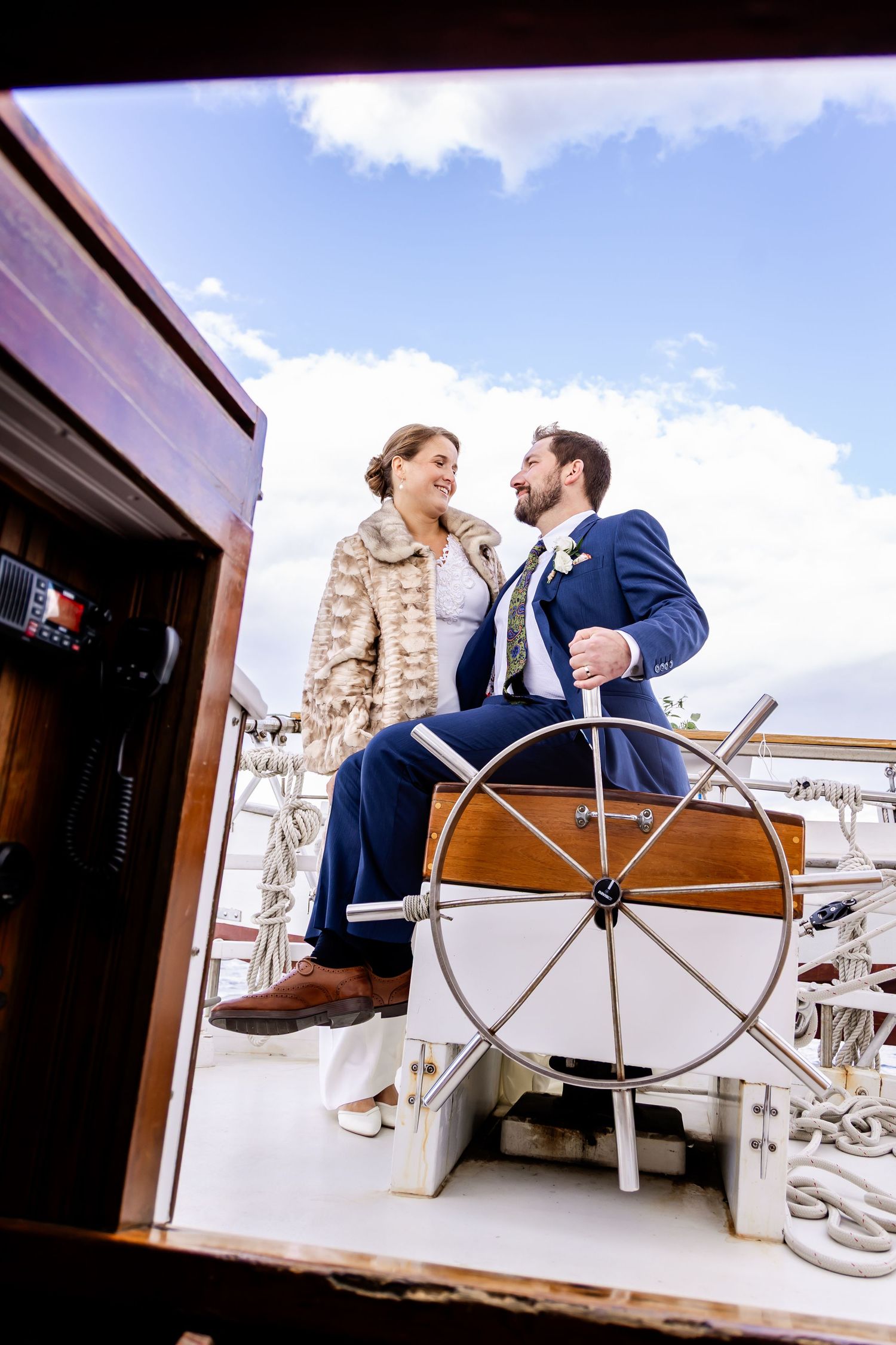 Bride and groom smiling at each other beside a ship's wheel on a sailboat deck under a blue sky.