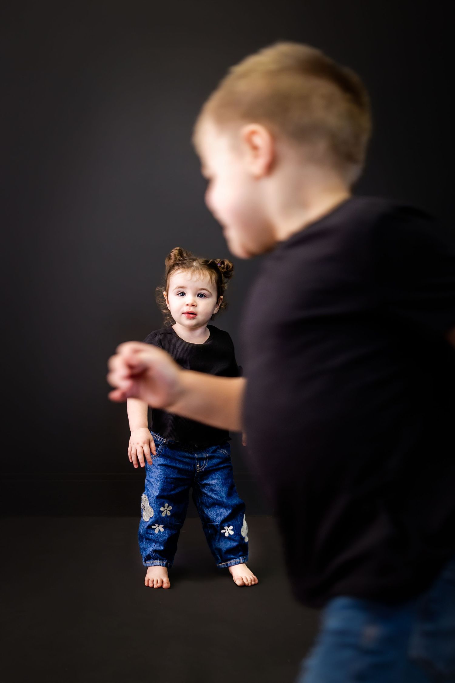 A barefoot toddler in a black shirt and flower-patterned jeans stands in a dark studio while a blurred child moves in the foreground.