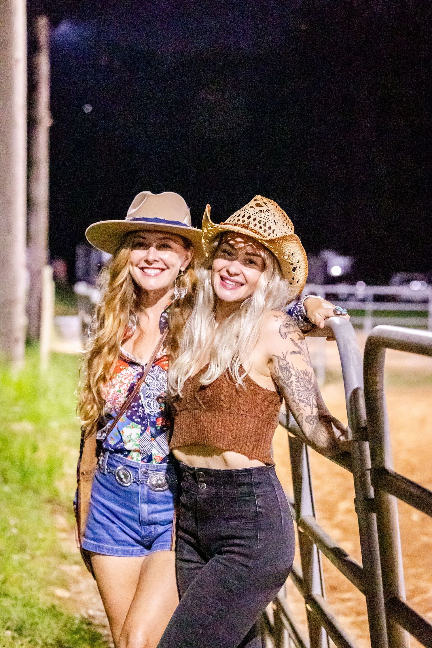 Two friends in cowboy hats lean against a fence at night wearing western-inspired outfits.