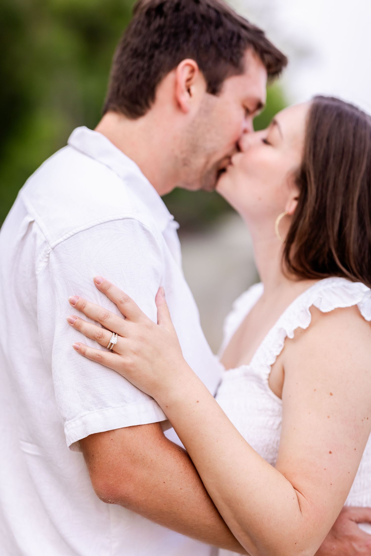 A tender romantic moment between a couple sharing a kiss outdoors, with the woman wearing white and displaying her ring.