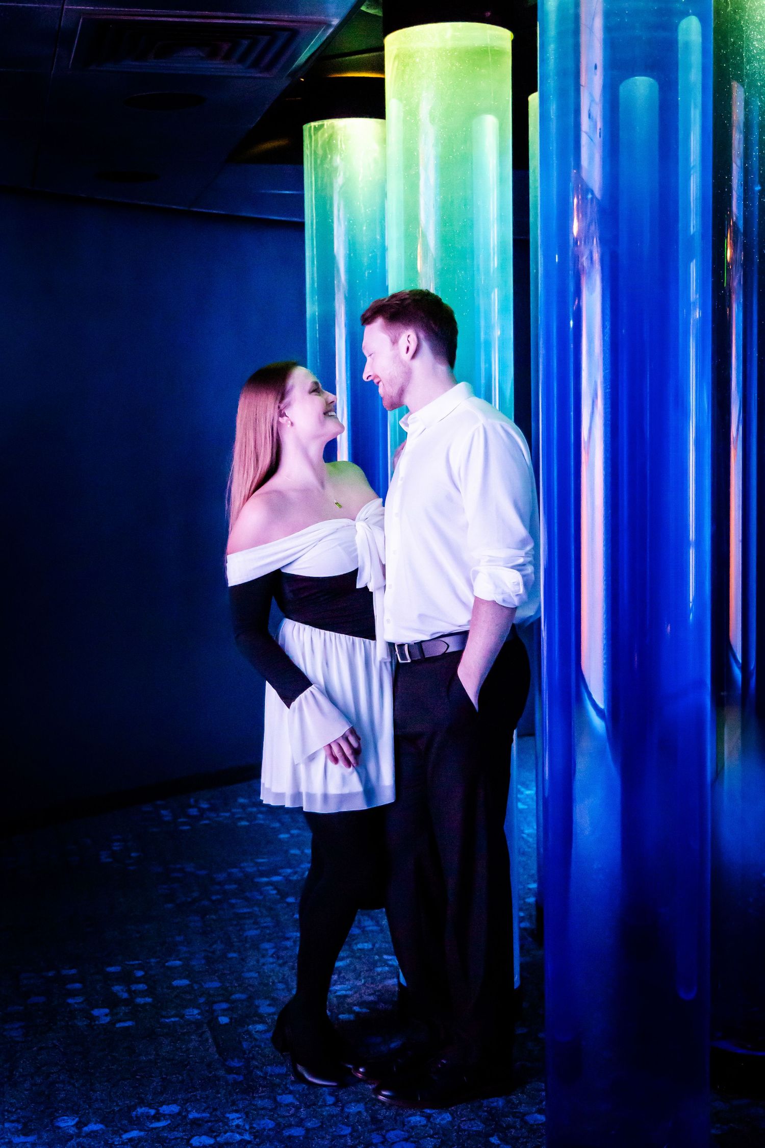 A romantic couple shares an intimate moment by glowing blue and green illuminated columns in a dark space.