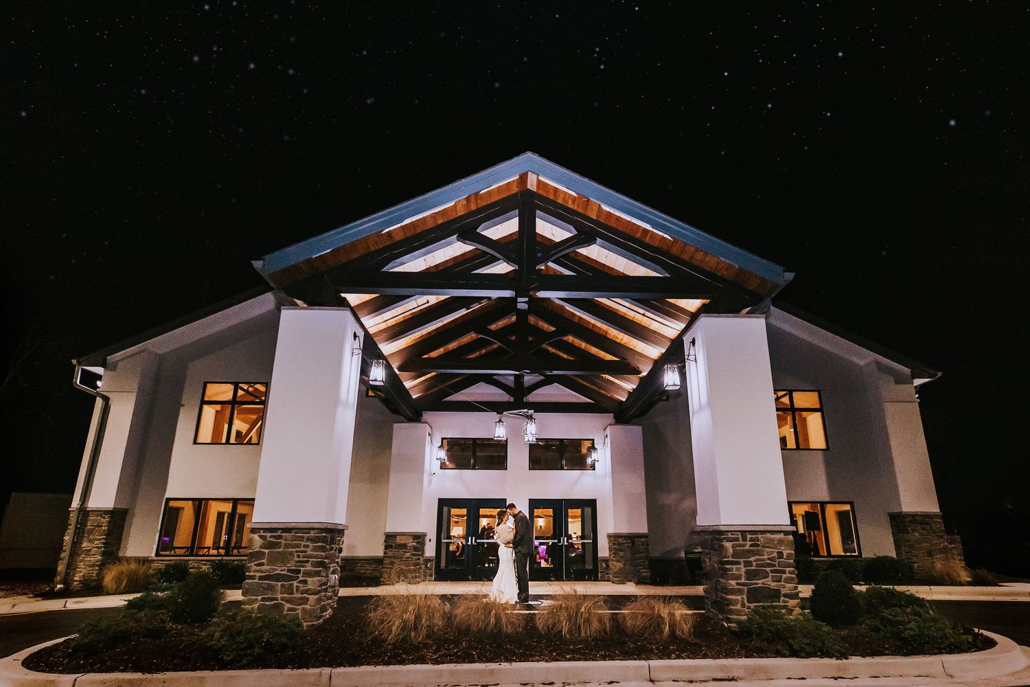 Modern white wedding venue with exposed wooden beams illuminated at night against a dark sky.