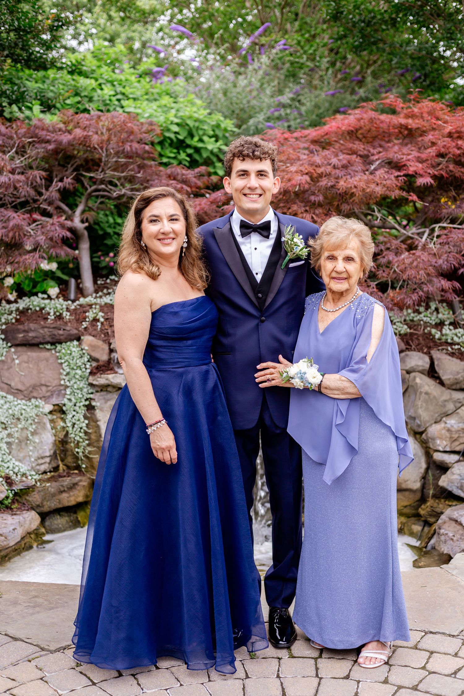 Wedding party members in navy blue and lavender formal attire pose together in a garden setting with red maple trees.