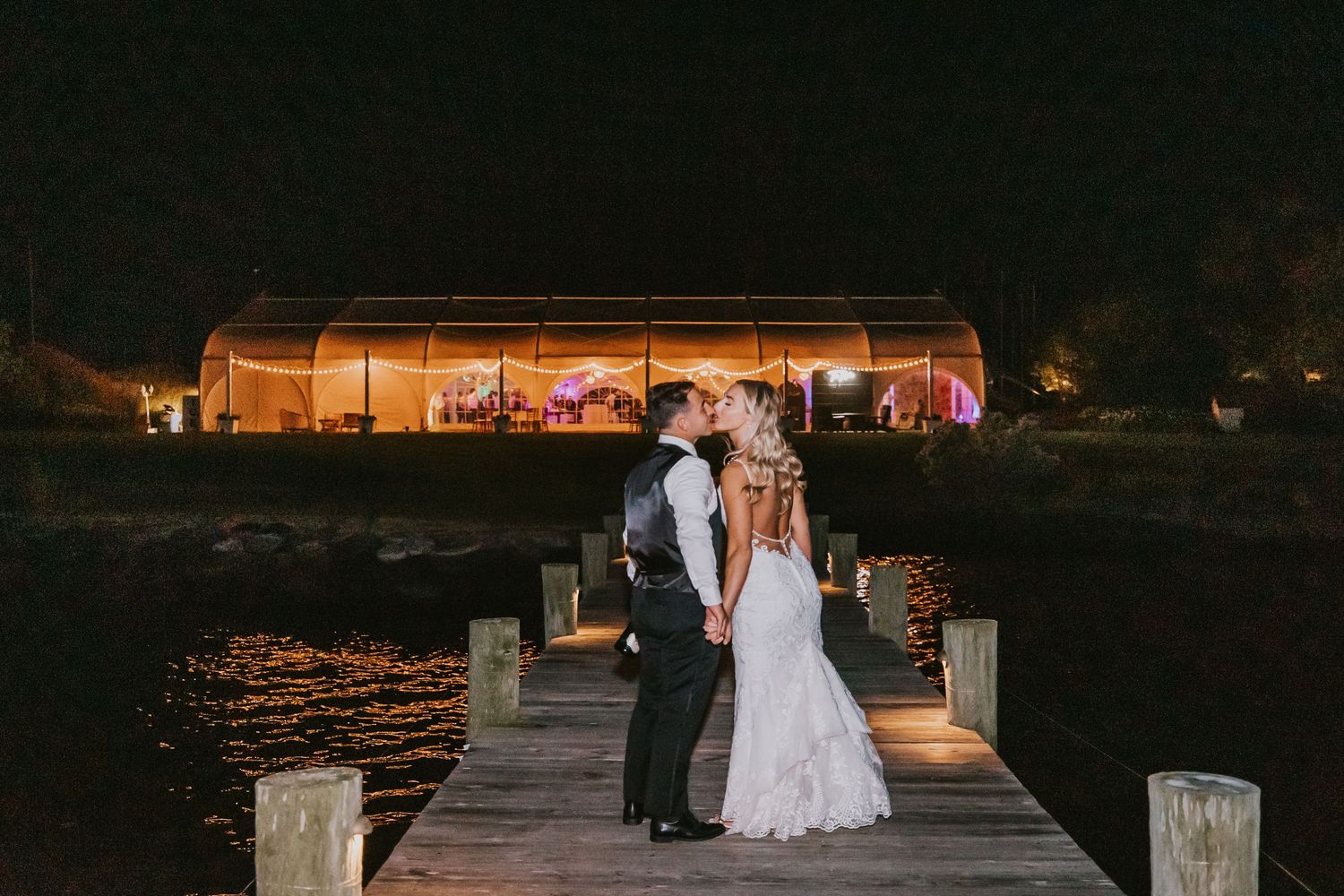 A bride and groom pose on a dock at night with an illuminated reception tent behind them.