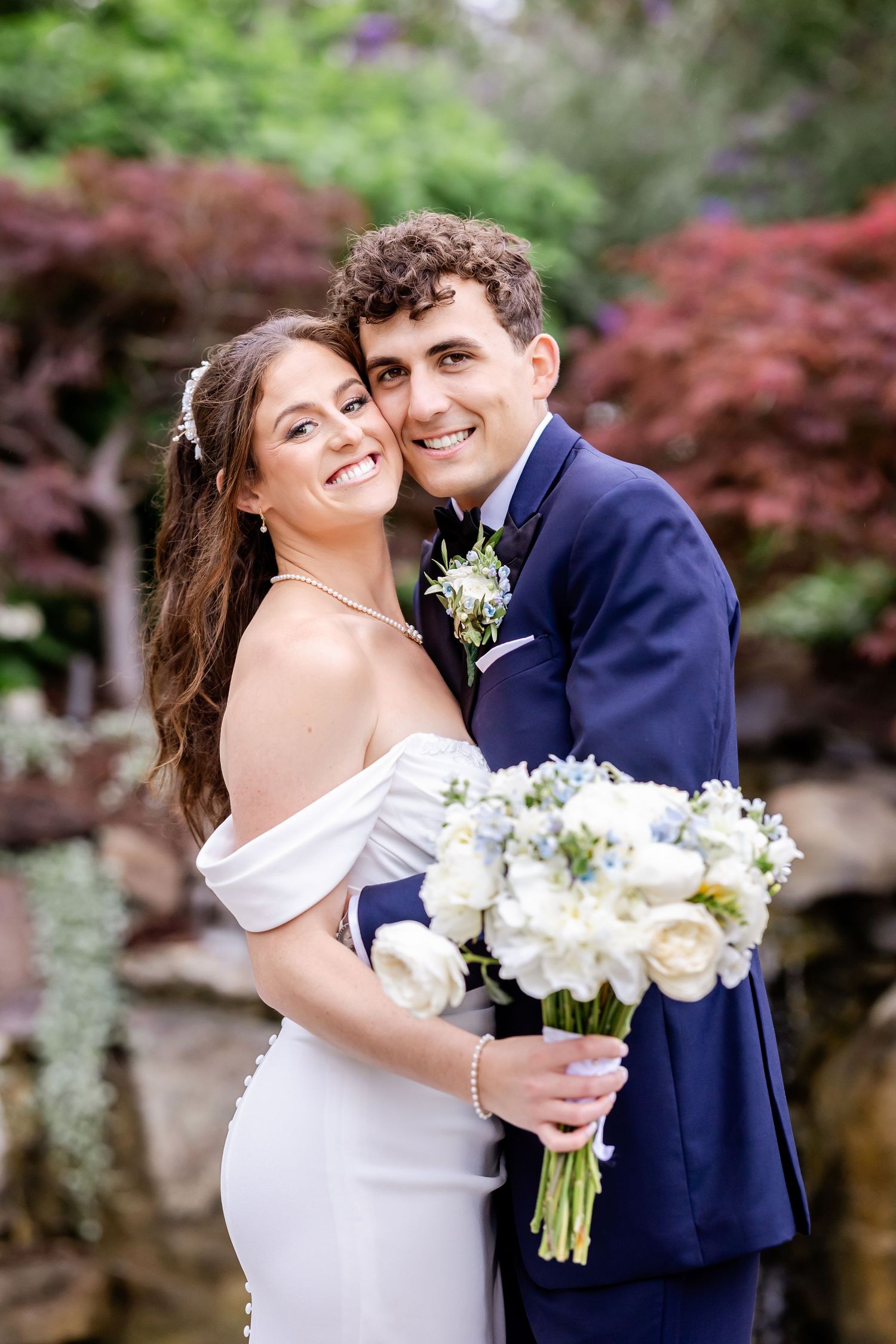 A joyful couple on their wedding day poses outdoors with a white bouquet against a backdrop of red maple trees.