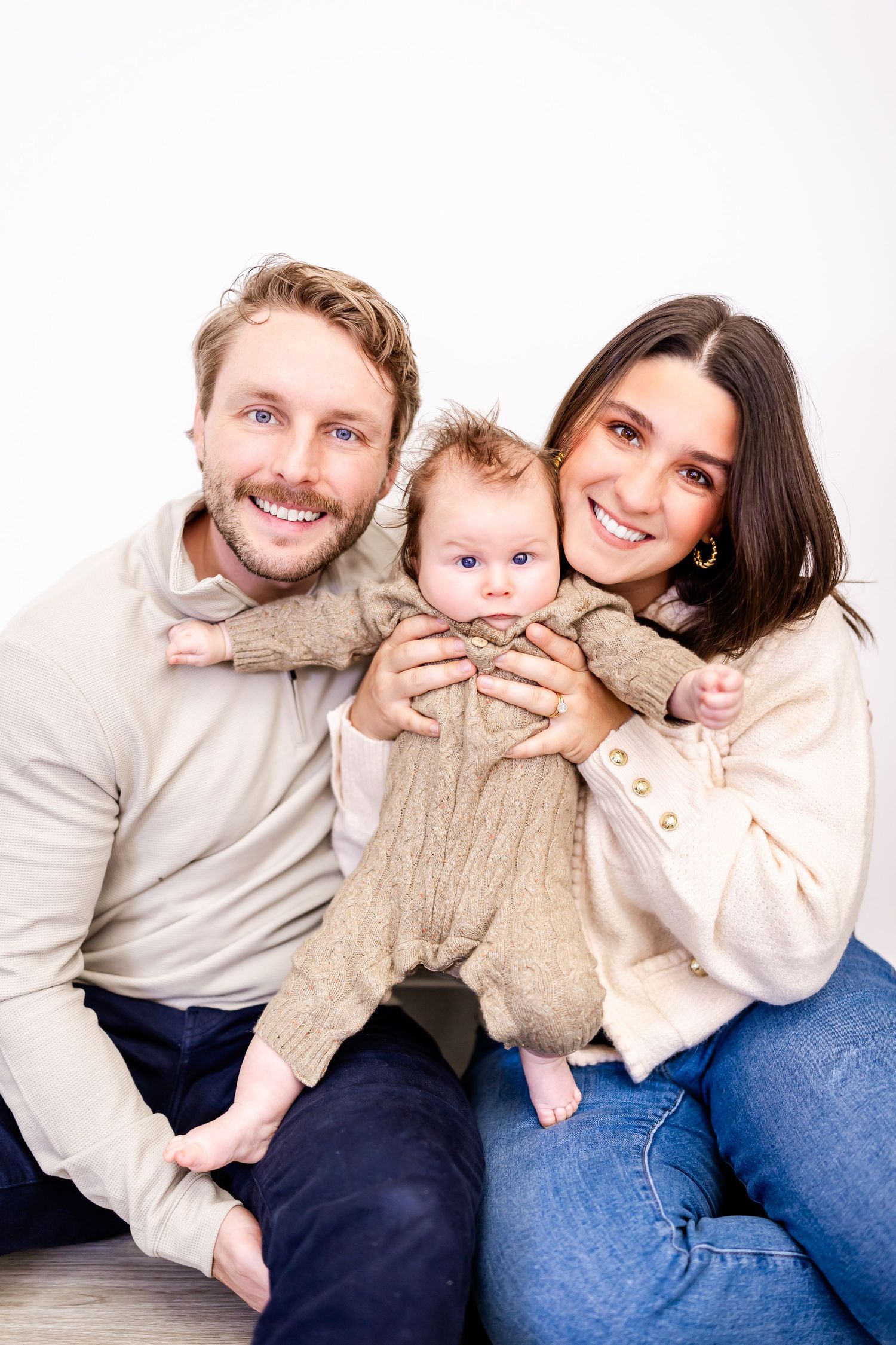 A happy family with a young baby wearing beige outfits poses together on a light background for a portrait.