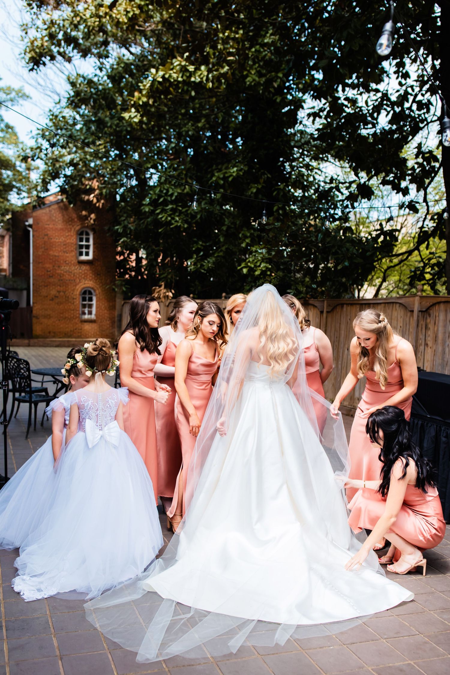Bridesmaids in coral dresses gather around bride in white wedding gown on outdoor stone patio during sunny celebration.