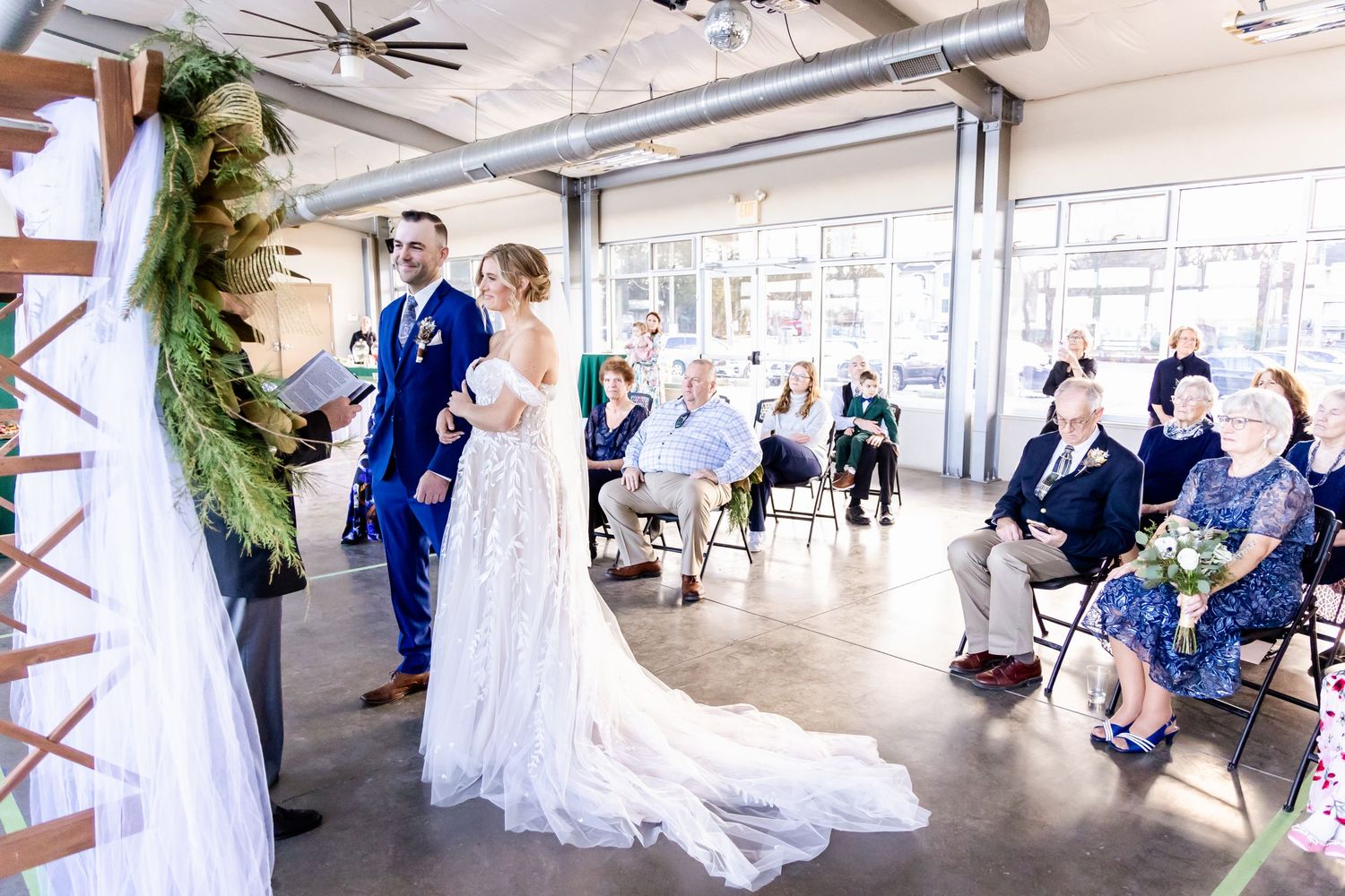 Wedding ceremony in an industrial venue with guests seated and greenery arch decor.