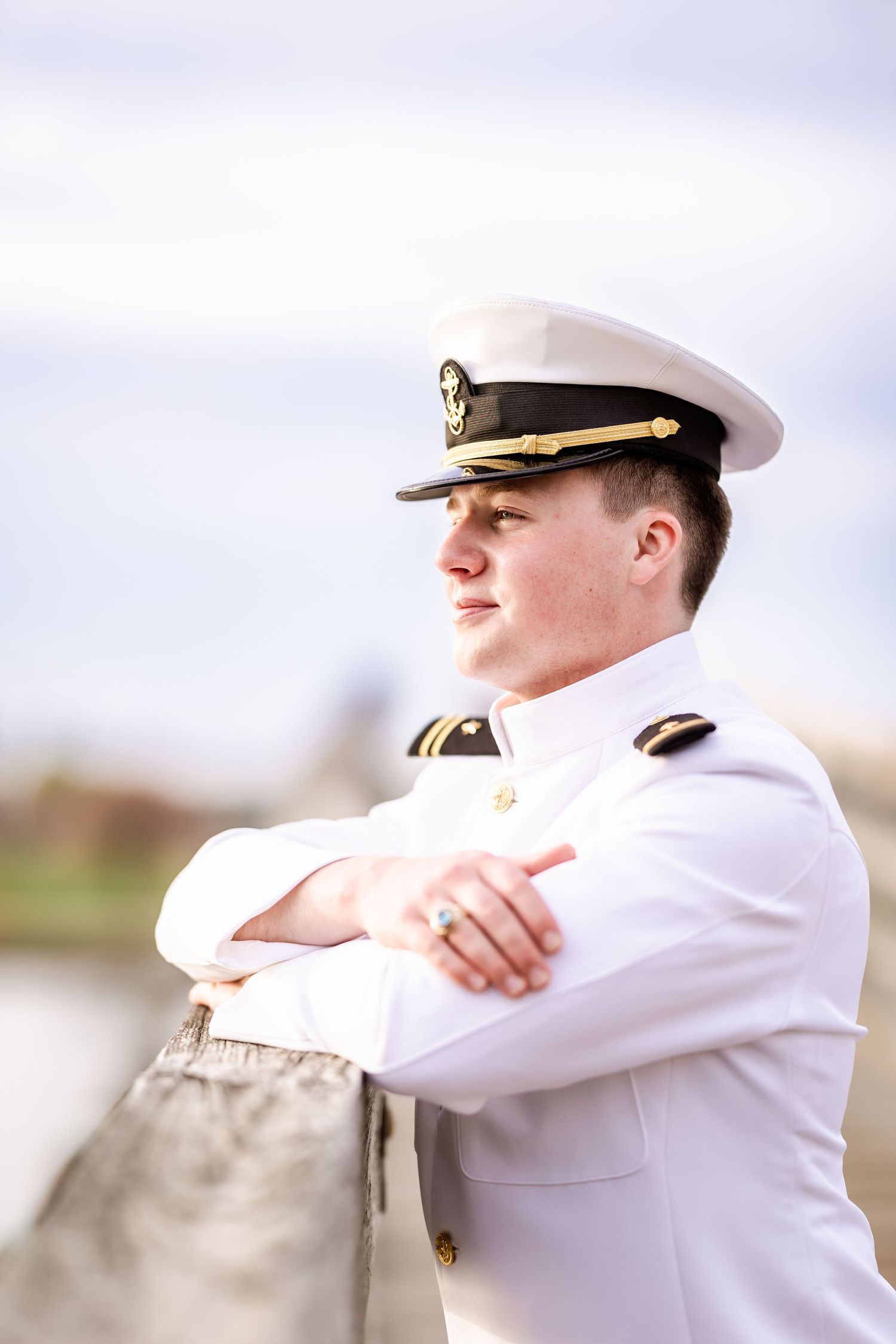 Profile view of naval officer in white dress uniform and cap leaning against railing outdoors.