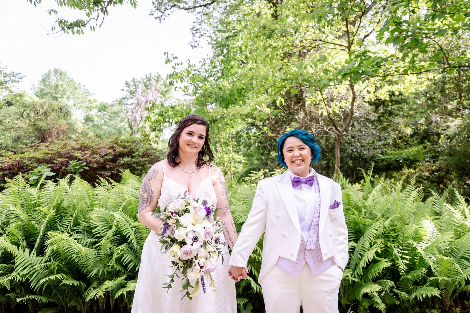 A wedding couple poses in a lush garden setting with ferns and greenery, one in white dress holding a floral bouquet.
