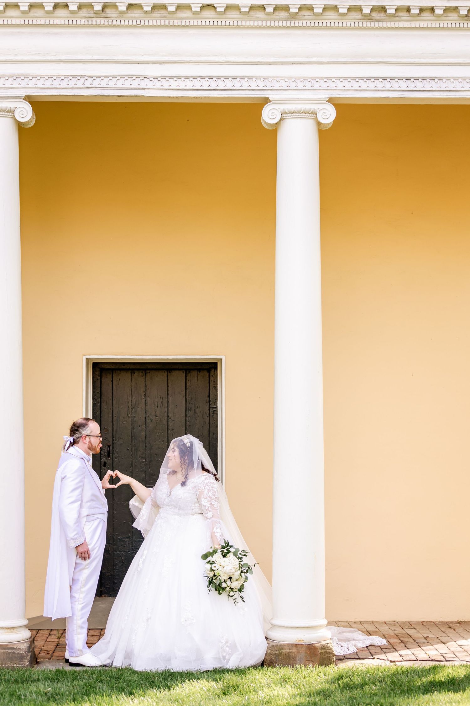 Wedding portrait with couple standing between grand white columns against yellow wall.