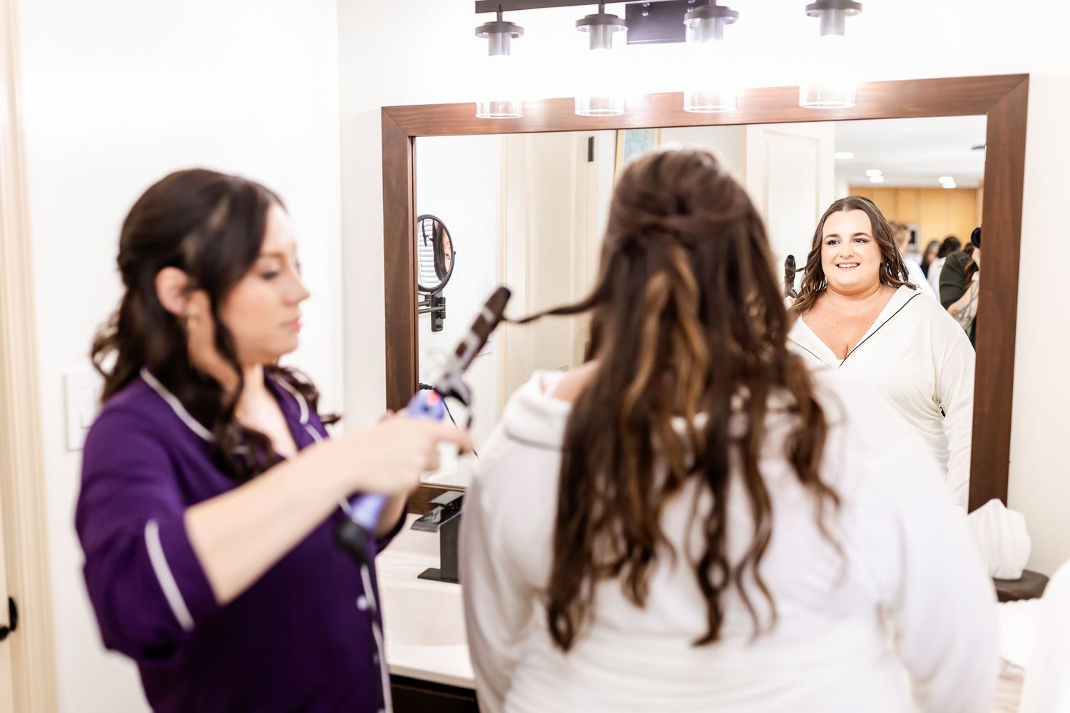 Hair and makeup artists work on styling in front of a brightly lit mirror.