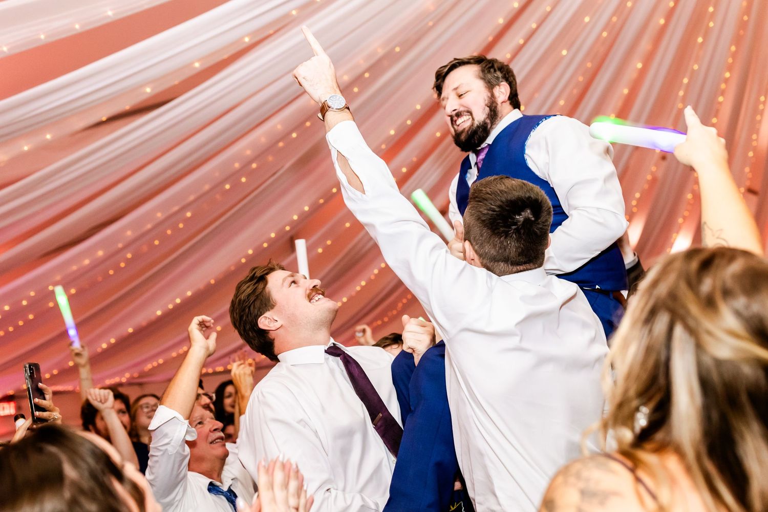 Celebratory dance moves at a wedding reception under coral-colored tent draping.